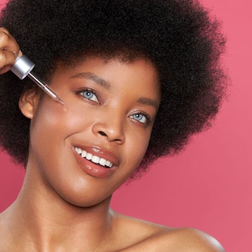 Cosmetics photo in pink background with an afro american model applying face serum on her cheek beauty product photography