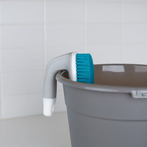 Product photography of a blue scrub brush with white and gray handle hanged on a gray bucket.