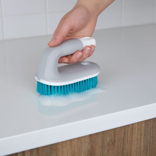 Female hand model holding a blue colored scrub brush with gray and white handle.