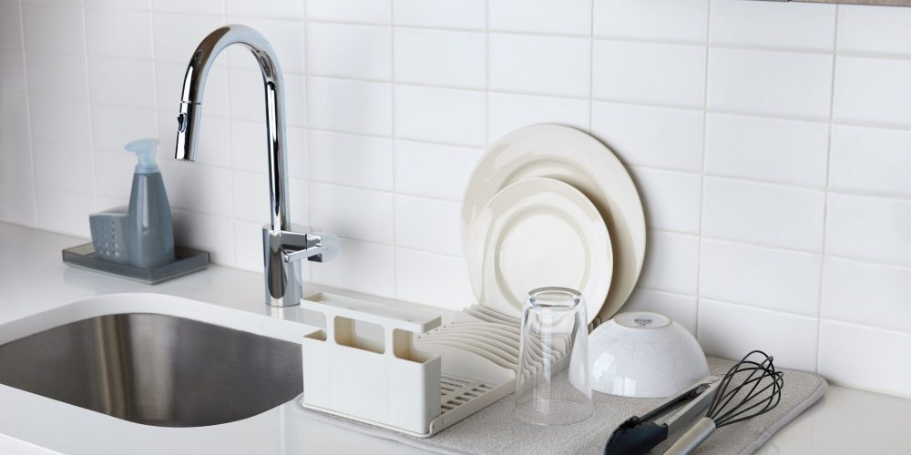 Lifestyle photography of kitchen accessories including dish tray, glass, bowl, plates and egg beater placed alongside of a sink.