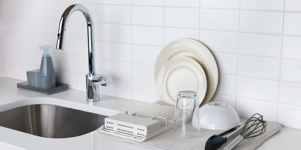 Lifestyle photography of kitchen accessories including dish tray, glass, bowl, plates and egg beater placed alongside of a sink.