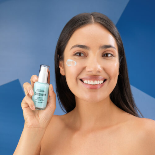 Female model holding a beauty product with a swatch placed on her face shot on a blue colored background.