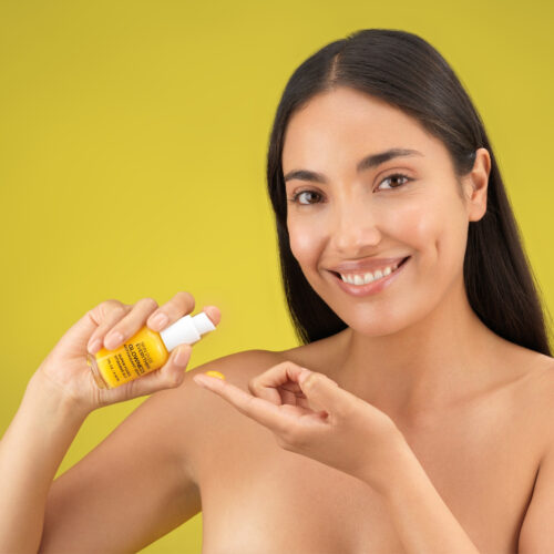 Cosmetics photography with female model holding a beauty product and pouring it on her finger shot on a yellow background.