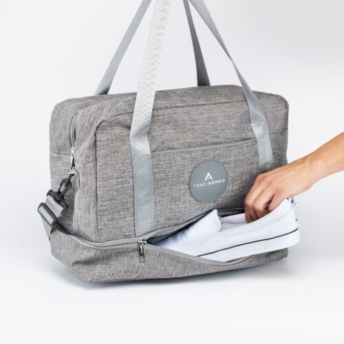 Female hand model putting white sneakers in the shoes compartment of a lucid grey colored travel bag.