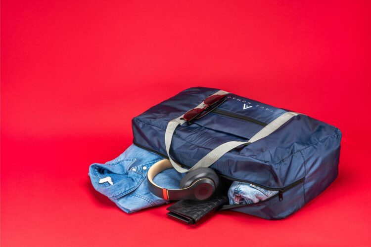 Creative lifestyle shot of a half opened travel bag in blue color revealing the items of the bag including shirt, headphones and purse on a red background.