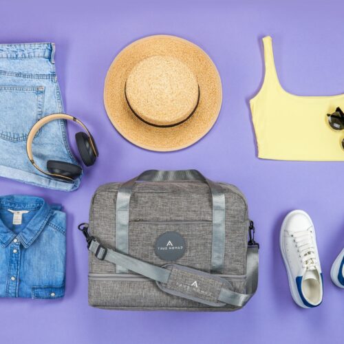 Creative lifestyle photography of a travel bag with necessary travelling items like hat, sunglasses, trouser, shirt, blouse and shoes on a purple background.