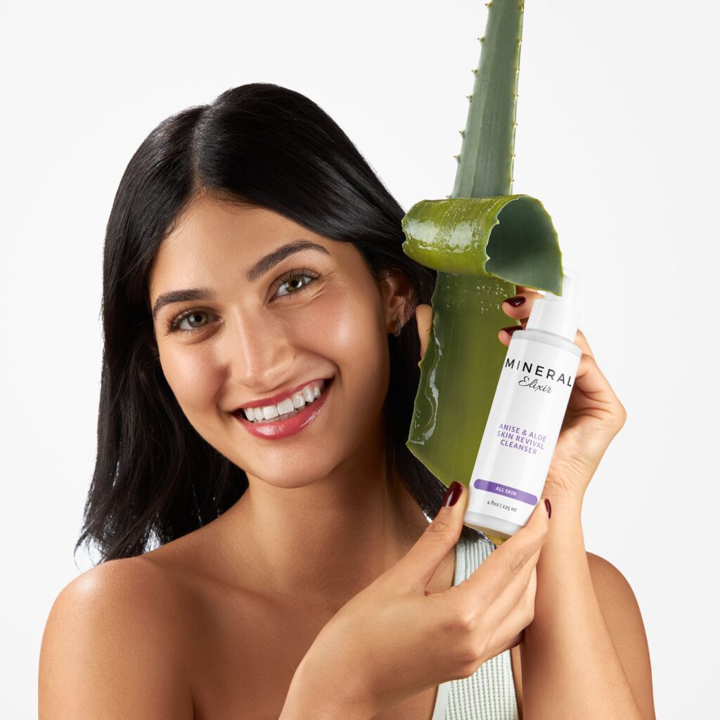 Cheerful woman holding aloe vera and a bottle of Mineral Enriched Aloe Skin Revivable Cleanser, highlighting organic skincare ingredients against a white backdrop.